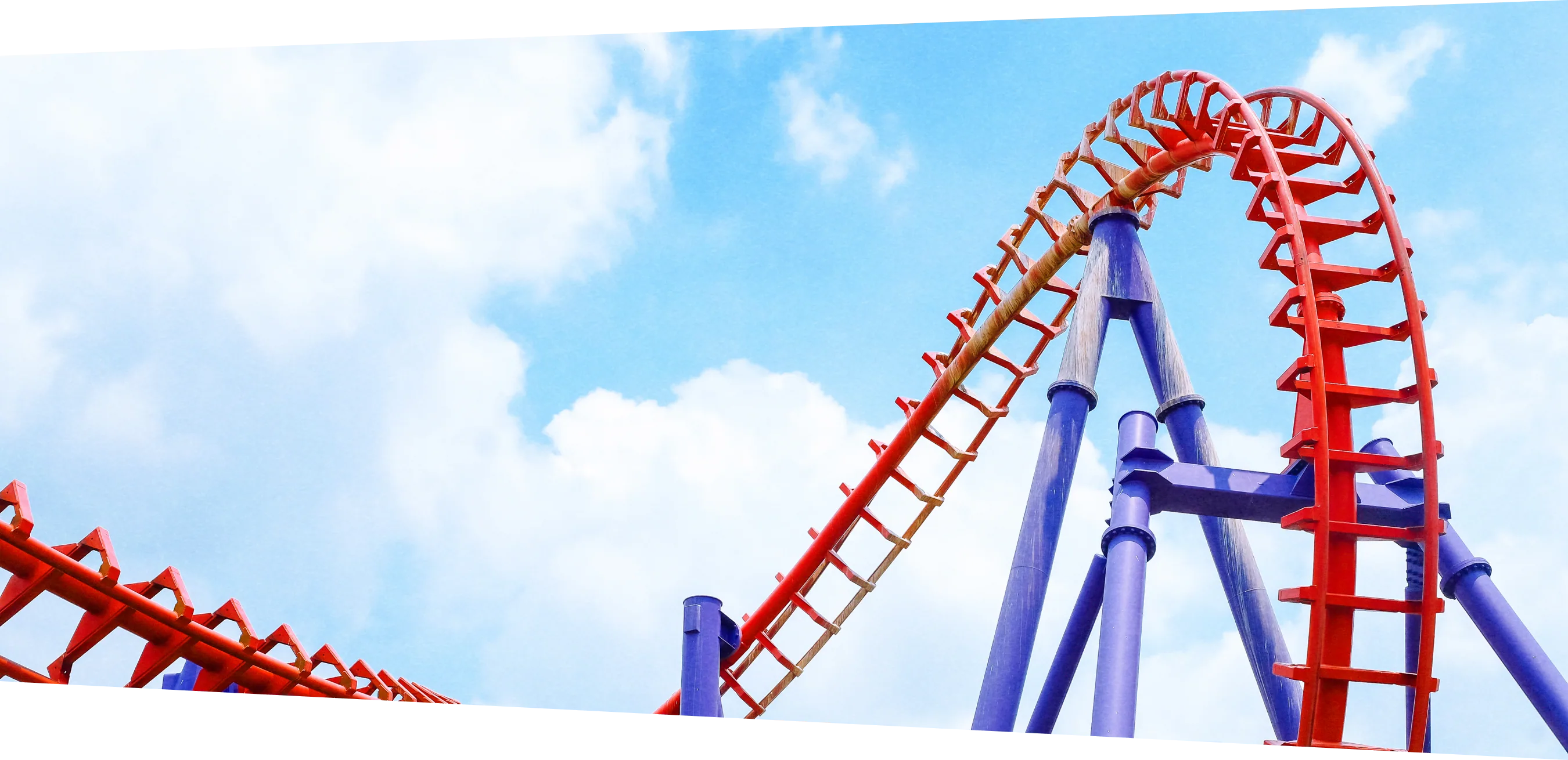 A red rollercoaster climbs into a bright, cloud-filled sky.